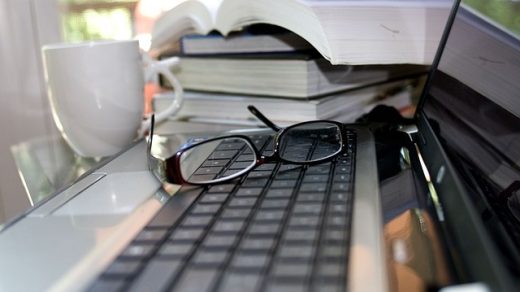 Kompuetr na którym leżą okulary a obok jest stos książek